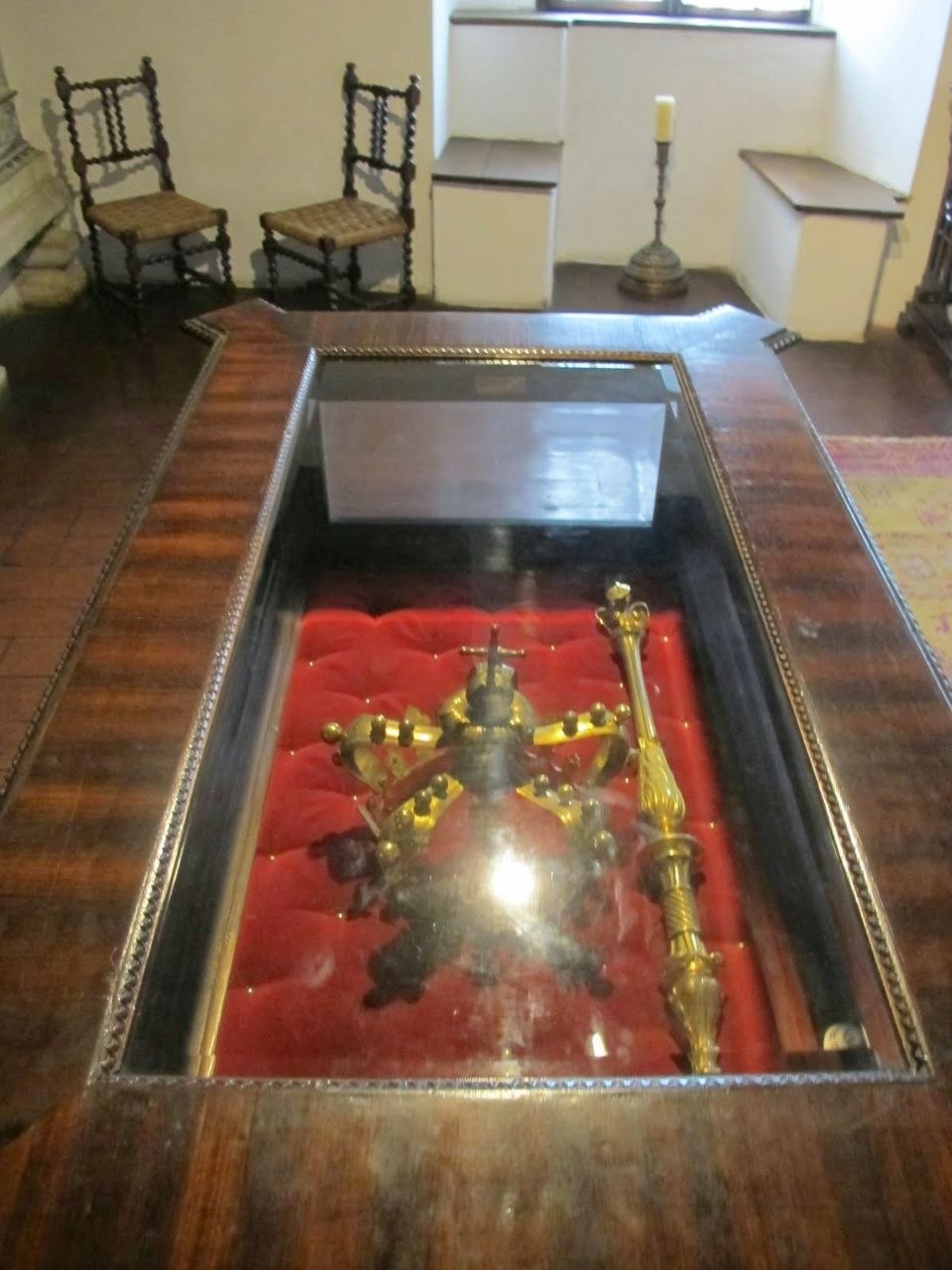 A royal crown and sceptre displayed in a glass case on a red velvet cushion inside Bran Castle — a dark metal crown with gilded orb and cross finial and decorative bosses, alongside a long gilded sceptre with an ornate top, viewed from above through the glass, with two small rush-seated chairs and a candlestick visible in the room behind